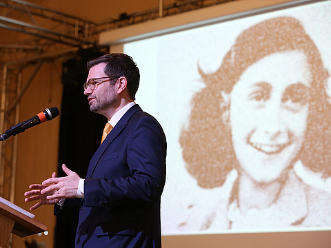 Ein Mann steht an einem Rednerpult mit einem Mikrofon und hält eine Rede. Im Hintergrund ist ein großes schwarz-weiß Porträt eines jungen Mädchens an die Wand projiziert.