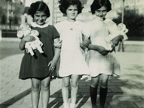 Drei Mädchen stehen nebeneinander auf einem Bürgersteig. Zwei von ihnen halten Puppen im Arm. Im Hintergrund stehen ein Hochhaus und kleinere Häuser daneben.