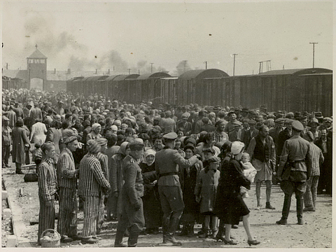 Eine große Menschenmasse steht dicht gedrängt beisammen. Im Vordergrund stehen Männer in Uniform und in gestreifter Kleidung. Im Hintergrund steht ein langer Güterzug und ein Tor.