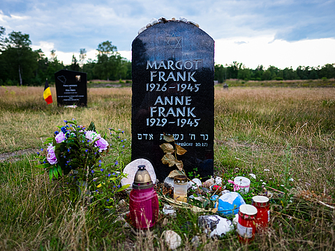 Ein schwarzer Grabstein mit weißer Schrift steht auf einer Wiese. Vor dem Stein befinden sich Kerzen, daneben steht ein Blumenstrauß.