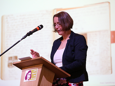 Eine Frau mit kurzen braunen Haaren steht an einem Rednerpult. Sie liest von einem Zettel ab und spricht in ein Mikrofon.