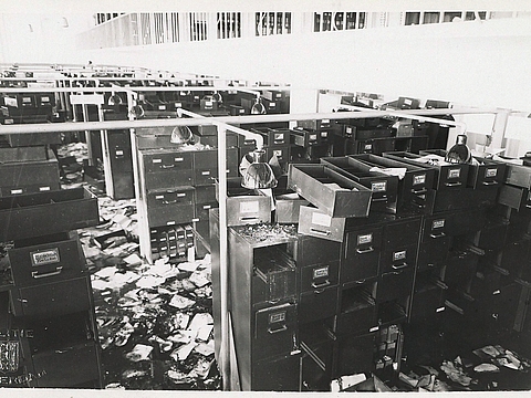 In Reihen stehende Aktenschränke in einem großen Raum. Viele Schubladen befinden sich außerhalb der Schränke, der Boden ist von losem Papier bedeckt.