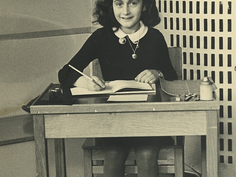 Eine lächelnde Jugendliche sitzt an einem Pult mit einem Stift in der Hand. Auf dem Tisch liegt ein aufgeklapptes Buch.