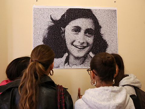 An einer Wand hängt ein schwarz-weiß Porträt eines jungen, lächelndes Mädchens. Vier Schülerinnen stehen mit dem Rücken zur Kamera und betrachten das Porträt.