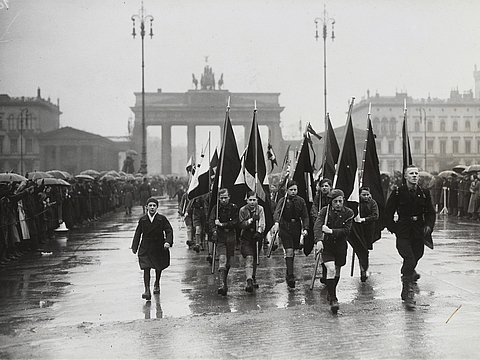 Eine Gruppe männlicher Jugendlicher in Uniform läuft in Formation und trägt Flaggen. Im Hintergrund das Brandenburger Tor in Berlin.