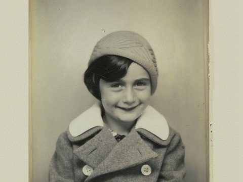 Ein lächelndes Mädchen sitzend in einem Fotoautomaten. Um das Foto herum ist ein Rahmen aus Papier, der das Gewicht des Mädchens, das Datum der Aufnahme und den Aufnahmeort angibt.