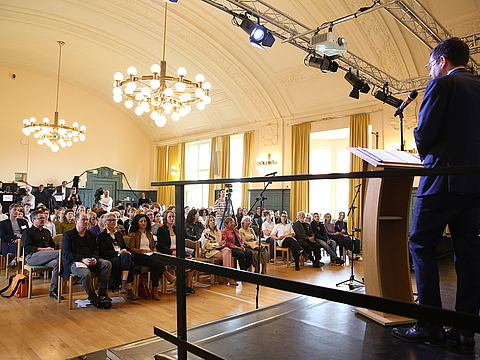 In einem großen Saal steht ein Mann auf einer Bühne an einem Pult mit einem Mikrofon und hält eine Rede. Im Hintergrund sitzen viele Menschen im Publikum, die dem Mann zu hören.