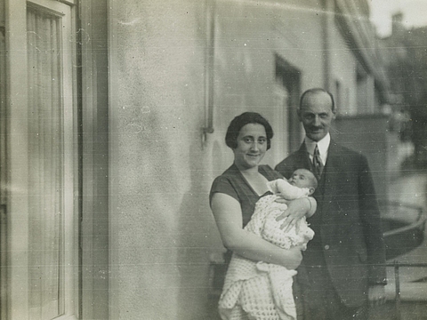 Eine Frau und ein Mann stehen vor einer Häuserwand. Die Frau hält ein Baby auf dem Arm.